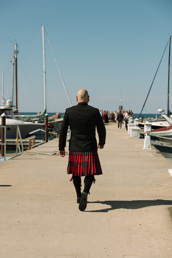 Groom walking in his kilt down dock toward sailboat for his wedding ceremony