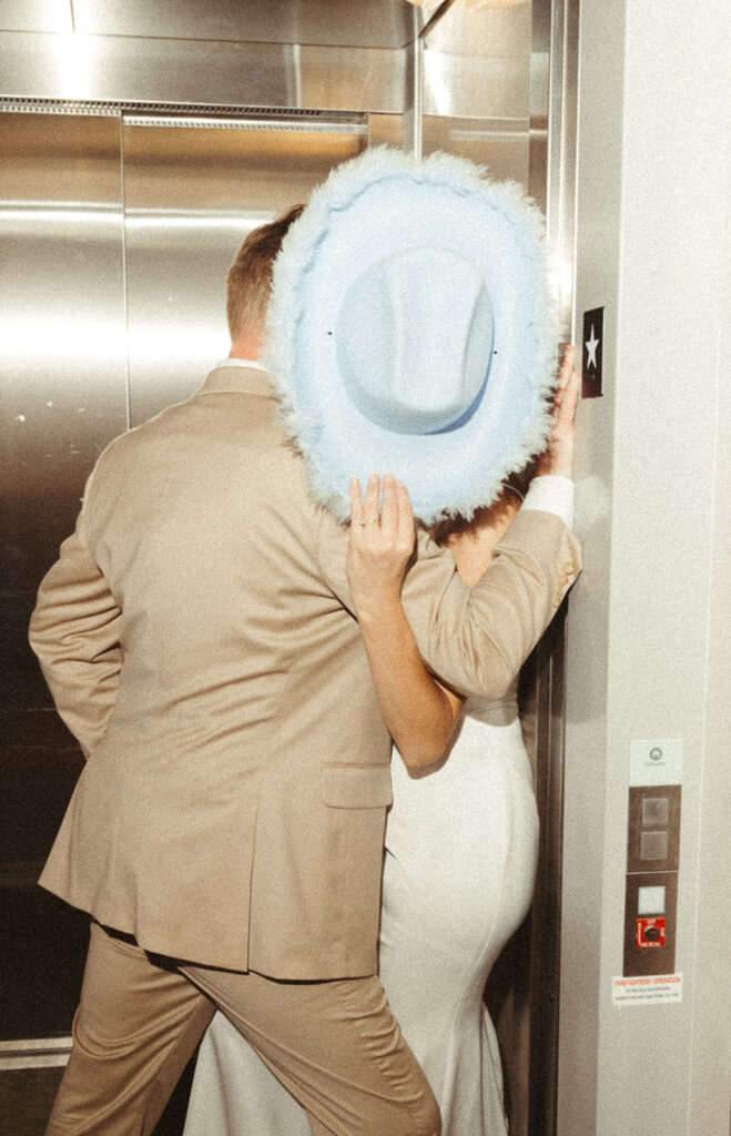 Couple in elevator kissing, while bride holds up cowgirl hat to block faces at Door County wedding reception