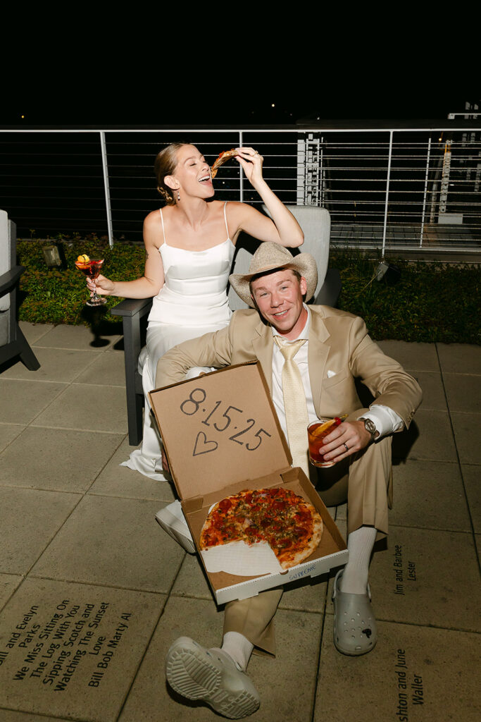 Groom holding a pizza box and bride eating pizza during a fun, non-traditional Door County wedding reception