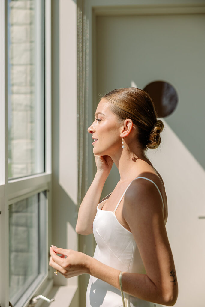 Bride adjusting her earrings in window light during a relaxed Door County wedding morning